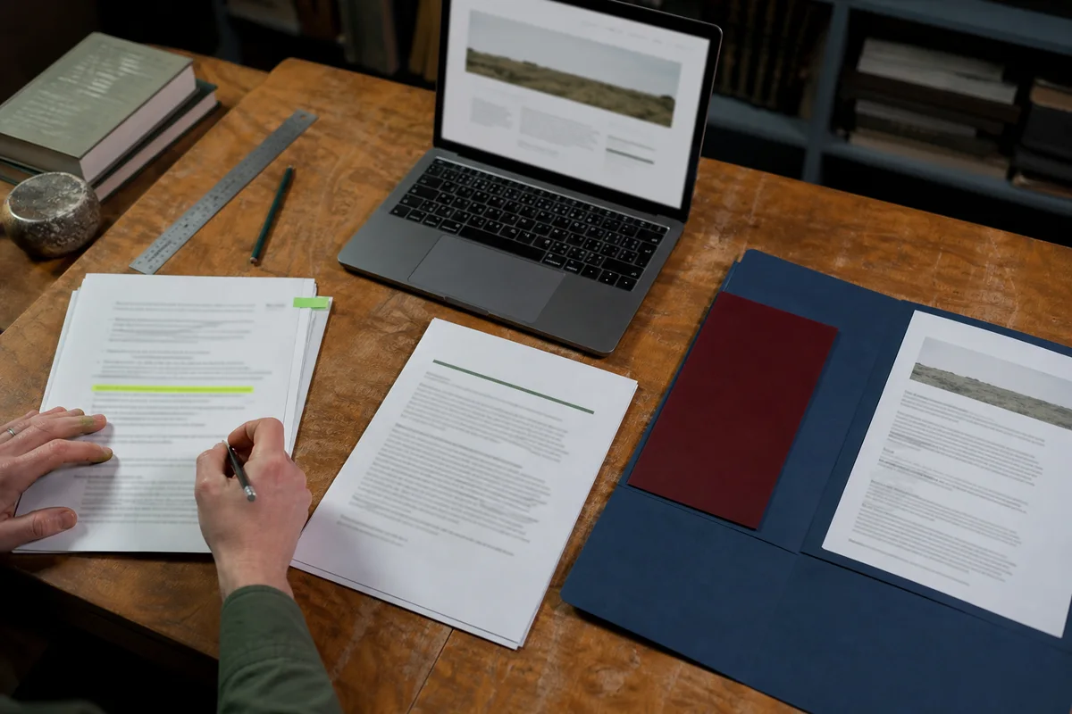 Editorial photograph of hands preparing a grant proposal beside scope sheets, a blue folder and a website preview
